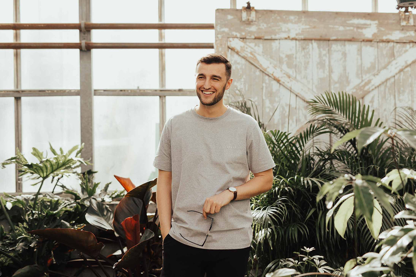 Smiling man holding his glasses in a grey t-shirt and standing in a greenhouse